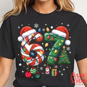 May include: Black t-shirt featuring the numbers "67" designed with Christmas elements. The "6" is candy cane-striped and the "7" is green with a Christmas tree. Both numbers are adorned with Santa hats and string lights.