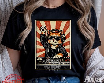 Puede incluir: Camiseta negra con un diseño gráfico de un gato DJ con gafas de sol y auriculares, con una pata levantada. El gato está detrás de una mesa de mezclas de DJ, sobre un fondo retro rojo y crema. También se ve el texto "Vibe Studio" y "Aya Vibe".