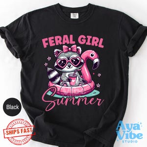May include: Black t-shirt with a graphic of a raccoon wearing heart-shaped sunglasses and a pink bow, floating on a flamingo float. The text "FERAL GIRL SUMMER" is displayed in pink script.