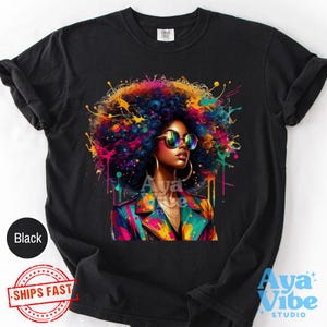 Könnte beinhalten: Ein schwarzes Kurzarm-T-Shirt mit einem lebendigen Grafikdruck einer Person mit einer großen, bunten Afro-Frisur, die eine runde Sonnenbrille und Creolen trägt. Das Design enthält helle Farbspritzer in Pink, Blau und Gelb.