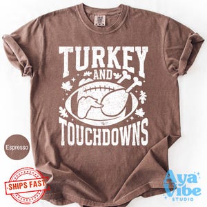 May include: A brown short-sleeved t-shirt featuring a white graphic print with the text "TURKEY AND TOUCHDOWNS". The design includes a football, a turkey leg, and autumn leaves. The shirt has a crew neckline and a relaxed, casual fit.