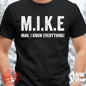 May include: Black t-shirt with the text "M.I.K.E. MAN, I KNOW EVERYTHING!" in white, distressed lettering. The shirt is a crew neck style and appears to be made of a soft material. The shirt is a casual wear item.
