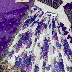 May include: A three-piece ensemble featuring a white skirt with a purple floral print and gold embroidery, a purple striped sleeveless crop top with thin straps, and a sheer purple dupatta with a silver border and the text SAREESUTRAIN.
