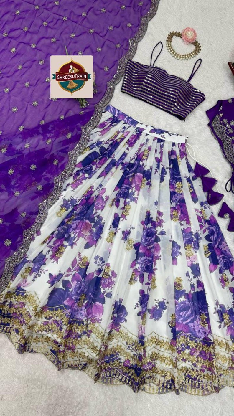 May include: A three-piece ensemble featuring a white skirt with a purple floral print and gold embroidery, a purple striped sleeveless crop top with thin straps, and a sheer purple dupatta with a silver border and the text SAREESUTRAIN.