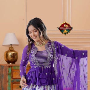 May include: A purple peplum top with long sleeves, silver embroidery, and a striped bodice paired with a white and purple floral print skirt. A matching purple sheer dupatta with silver embellishments completes the ensemble, featuring the SAREESUTRAIN logo.