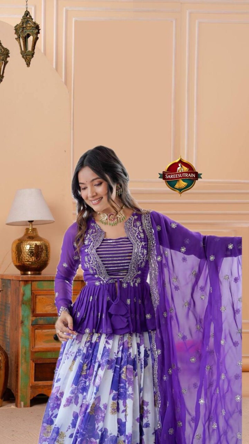 May include: A purple peplum top with long sleeves, silver embroidery, and a striped bodice paired with a white and purple floral print skirt. A matching purple sheer dupatta with silver embellishments completes the ensemble, featuring the SAREESUTRAIN logo.