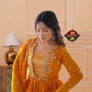 May include: A vibrant mustard yellow peplum-style top featuring long sleeves, a V-neckline, and intricate gold embroidery along the front edges. The bodice includes horizontal gold stripes, while the skirt displays a floral print in red and orange.
