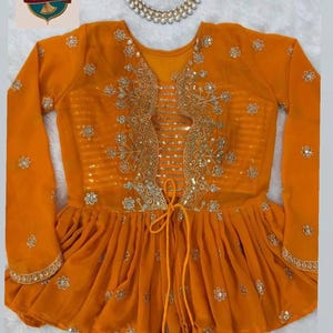 May include: A bright orange long-sleeved peplum blouse featuring intricate gold floral embroidery, a central panel with horizontal gold stripes, and a drawstring tie at the waist with tassel ends. A matching gold beaded necklace is positioned above.