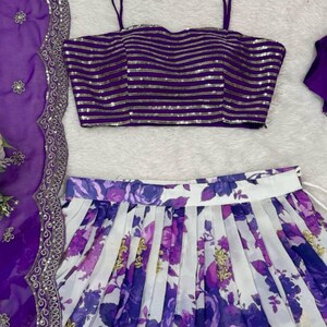 May include: A purple sleeveless crop top with horizontal gold sequin stripes, paired with a white pleated skirt featuring a purple floral print and gold accents, alongside a matching purple sheer dupatta with a decorative silver embroidered border.
