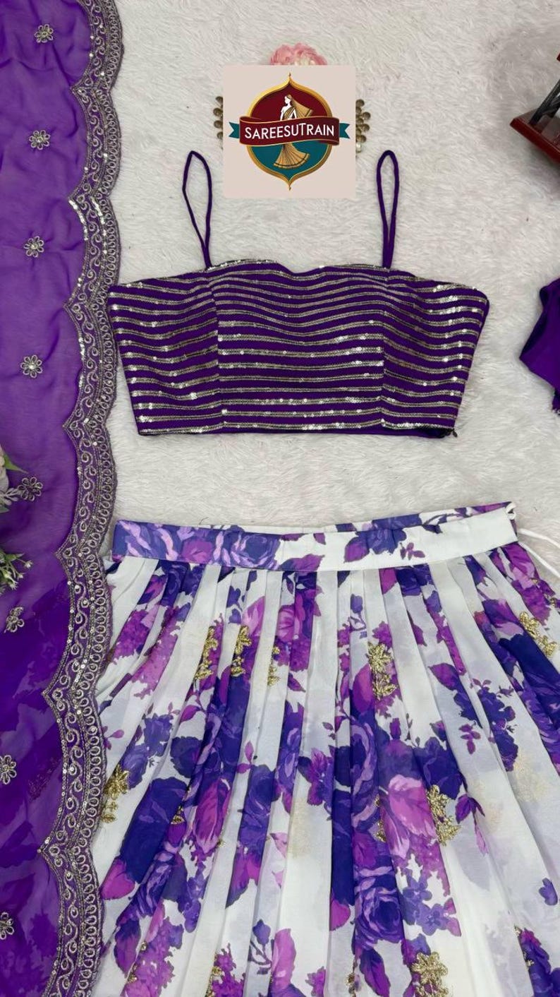 May include: A purple sleeveless crop top with horizontal gold sequin stripes, paired with a white pleated skirt featuring a purple floral print and gold accents, alongside a matching purple sheer dupatta with a decorative silver embroidered border.