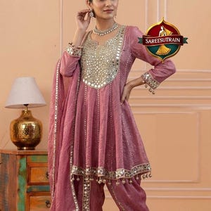 Puede incluir: Conjunto salwar kameez de color rosa empolvado. La túnica presenta intrincados trabajos de espejo y una silueta acampanada. Los pantalones a juego tienen un diseño único y fruncido. El atuendo se complementa con un dupatta y joyas a juego. El logotipo "SAREESUTRAIN" es visible.