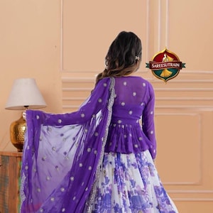 May include: A purple peplum top with long sleeves and small floral embellishments paired with a white and purple floral print skirt. A matching purple sheer dupatta with silver embroidery and small floral accents is draped over the shoulder.