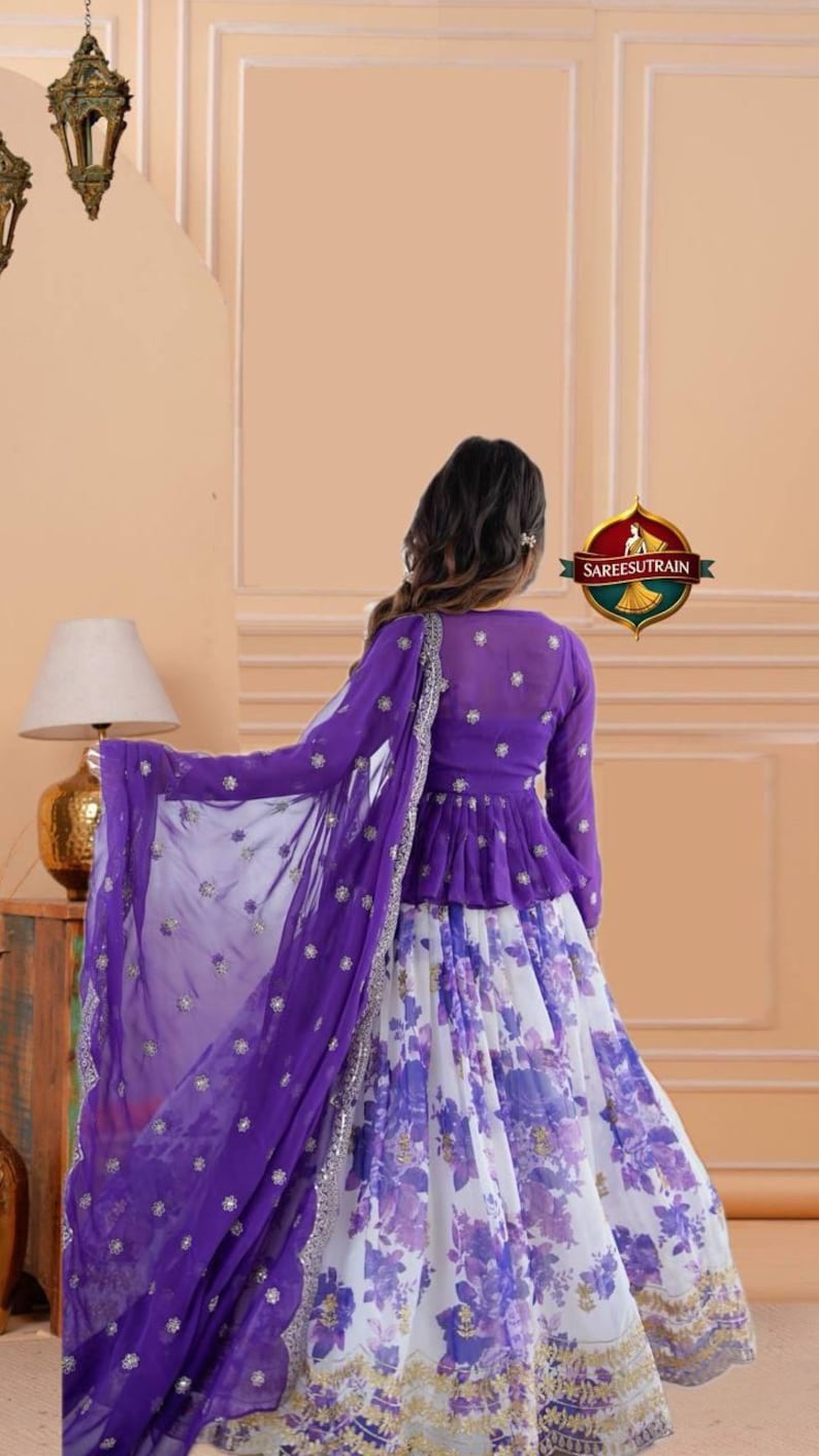 May include: A purple peplum top with long sleeves and small floral embellishments paired with a white and purple floral print skirt. A matching purple sheer dupatta with silver embroidery and small floral accents is draped over the shoulder.