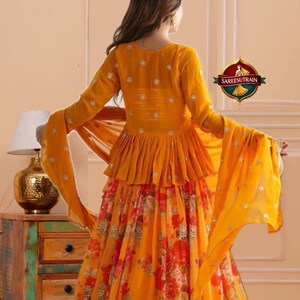 May include: A vibrant mustard yellow peplum top with long sleeves and silver embellishments paired with a matching floor-length skirt featuring a red and orange floral print and a gold embroidered hem. A sheer yellow dupatta is draped over the arms.