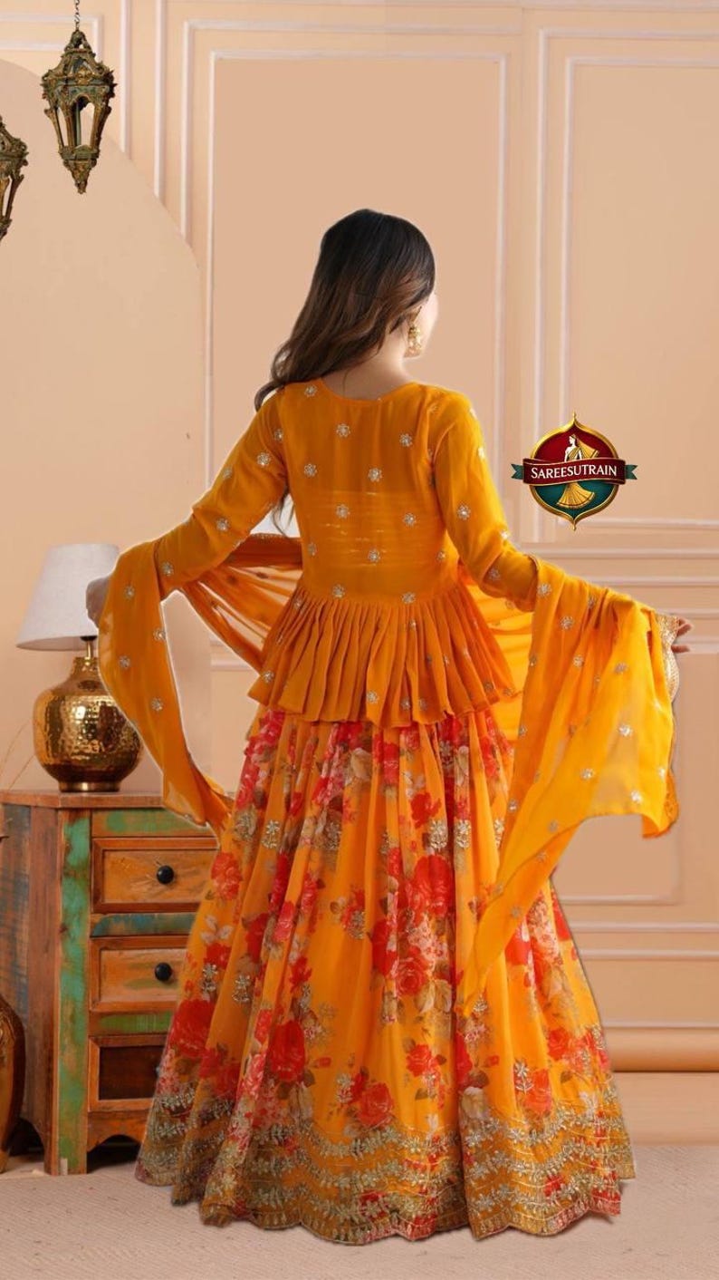 May include: A vibrant mustard yellow peplum top with long sleeves and silver embellishments paired with a matching floor-length skirt featuring a red and orange floral print and a gold embroidered hem. A sheer yellow dupatta is draped over the arms.