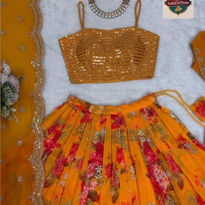 May include: A mustard yellow sleeveless crop top with horizontal gold sequin stripes, a pleated skirt featuring a vibrant red and orange floral print with scattered gold embellishments, and a matching sheer dupatta with a scalloped gold border.