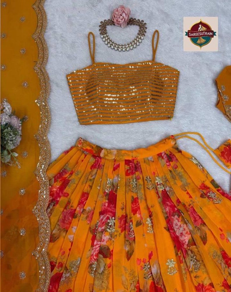 May include: A mustard yellow sleeveless crop top with horizontal gold sequin stripes, a pleated skirt featuring a vibrant red and orange floral print with scattered gold embellishments, and a matching sheer dupatta with a scalloped gold border.