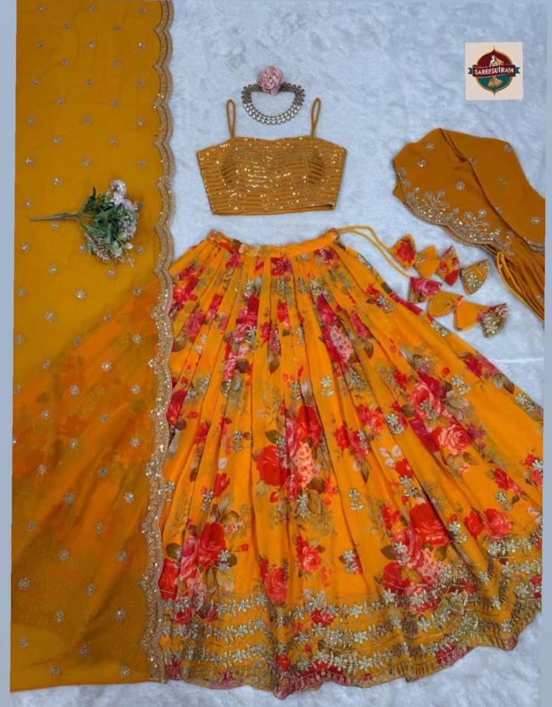 May include: A mustard yellow lehenga choli set featuring a sleeveless blouse with horizontal sequin stripes, a flared skirt with a vibrant red and orange floral print, and a matching dupatta with scalloped gold embroidered borders and small sequins.