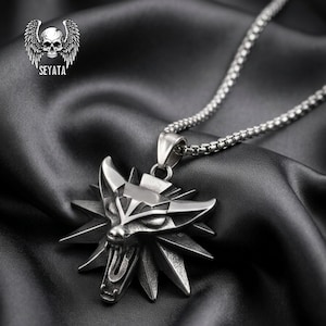 Puede incluir: Un collar con colgante plateado con un diseño de cabeza de lobo y un patrón geométrico de estrella. El colgante cuelga de una cadena a juego. La imagen también muestra un logotipo con una calavera y alas y el texto "SEYATA".
