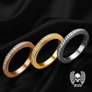 May include: Three rings in rose gold, gold, and gunmetal, each set with small, clear gemstones. The rings are displayed on a black satin surface. A logo with a skull and wings is in the bottom right corner.