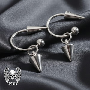 May include: Two silver-colored, curved barbell earrings with a conical spike at each end. Each earring has a small, dangling conical spike. The earrings are displayed on a dark gray, textured surface. The logo "SEYATA" is in the lower left corner.