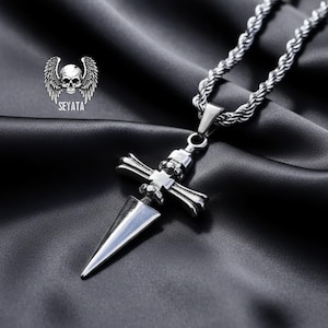 May include: A silver-colored pendant necklace featuring a dagger-shaped pendant with a cross-like design and a rope chain. The pendant has a skull and wing logo with the text "SEYATA". The necklace is displayed on a black satin background.