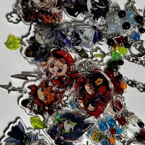 May include: A collection of anime-style character keychains. Each keychain features a colorful illustration of a character with detailed outfits and expressions. The keychains are attached to silver chains and adorned with beads and charms.
