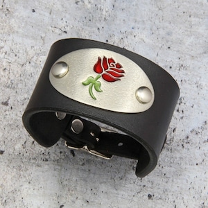 May include: A black leather cuff bracelet with a silver oval charm featuring a red rose with green leaves.