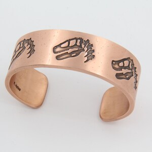 Puede incluir: Brazalete de puño de cobre con diseños de esqueletos de dinosaurios grabados. El diseño de puño abierto es de color cobre cálido y presenta tres imágenes de esqueletos de dinosaurios. El brazalete está estampado con el nombre "R. Hales".
