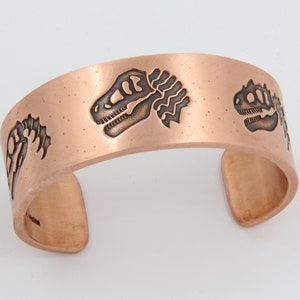 Puede incluir: Una pulsera de cobre con tres esqueletos de dinosaurios grabados. La pulsera tiene un diseño simple y redondeado.