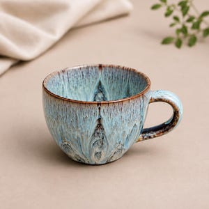 May include: A ceramic mug featuring a reactive glaze with a blend of light blue, white, and dark brown tones. The exterior and interior display a vertical, drip-like pattern with a smooth, rounded handle and a dark brown rim.