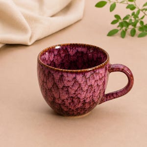 Può includere: Una tazza in ceramica con uno smalto reattivo color magenta intenso e viola scuro, caratterizzata da un motivo testurizzato simile a scaglie. La tazza presenta un corpo arrotondato, un'ampia apertura circolare e un manico curvo sul lato destro.