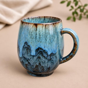 May include: A ceramic mug with a rounded body and a curved handle, featuring a reactive glaze finish that transitions from a light sky blue at the rim to a deep, dark navy blue at the base, with brown accents along the rim and handle edges.