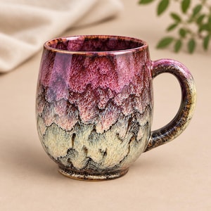 May include: A ceramic mug featuring a crystalline glaze with a gradient transition from deep magenta at the rim to a pale cream and dark brown base. The mug has a rounded body, a sturdy handle, and a textured, glossy finish with intricate patterns.