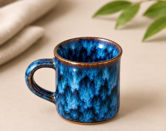 Può includere: Una tazza in ceramica con smalto reattivo blu profondo e nero con un motivo cristallino. La tazza presenta un manico arrotondato e una finitura liscia e lucida con un bordo marrone scuro che si estende fino alla base.