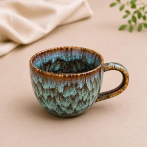 May include: A ceramic mug featuring a reactive glaze with a blend of deep brown and turquoise blue colours. The exterior and interior display a mottled, drip-like pattern that transitions from a dark brown rim to a vibrant, textured teal base.
