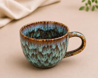 May include: A ceramic mug featuring a reactive glaze with a blend of deep brown and turquoise blue colours. The exterior and interior display a mottled, drip-like pattern that transitions from a dark brown rim to a vibrant, textured teal base.