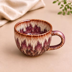 Può includere: Una tazza in ceramica con smalto reattivo caratterizzata da un motivo a forma di montagna nei toni del viola intenso e del crema. La tazza presenta un corpo arrotondato e un manico curvo, con lo smalto che cola dal bordo in un effetto materico.