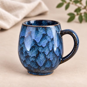 May include: A rounded ceramic mug featuring a deep blue and light blue mottled glaze with a scale-like pattern. The mug has a curved handle and a smooth, glossy finish, with dark brown edges visible along the rim and the base of the handle.