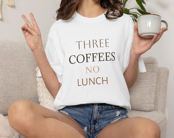 Puede incluir: Una camiseta blanca de manga corta con el texto "THREE COFFEES NO LUNCH" impreso en letras negras y marrones. La camiseta se combina con pantalones cortos de mezclilla azul y se sostiene una taza de cerámica de color claro en una mano.