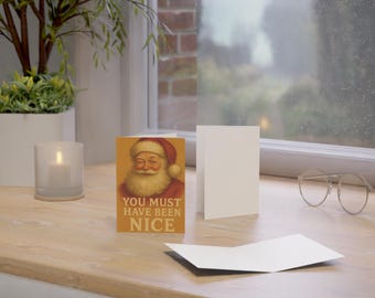 Può includere: Un biglietto di Natale con un'illustrazione vintage di Babbo Natale in abito e cappello rossi. Il biglietto recita "YOU MUST HAVE BEEN NICE". Ci sono due biglietti bianchi vuoti e una candela accesa in un bicchiere.