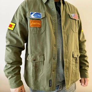 May include: Olive green shirt with multiple pockets and patches. Patches include the text "Fastend" and "Frontier", and a small American flag. The shirt has a casual, utilitarian style.