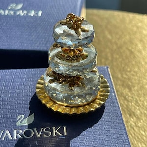 以下が含まれることがあります： 金色のアクセントが付いた、3段のクリスタルケーキの置物。透明なクリスタルの層には金色の花の装飾が施され、金色のベースの上に置かれています。置物は青い箱に飾られています。