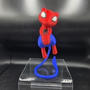 May include: A 3D-printed figurine of a cat-like character in a Spider-Man costume. The figure is primarily red with blue accents, featuring white eyes and a black spider emblem. A blue tail extends from the figure, resting on a clear display stand.