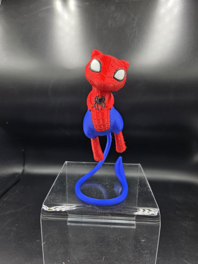May include: A 3D-printed figurine of a cat-like character in a Spider-Man costume. The figure is primarily red with blue accents, featuring white eyes and a black spider emblem. A blue tail extends from the figure, resting on a clear display stand.