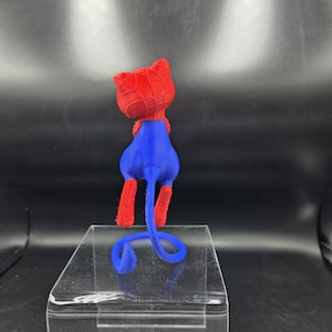 May include: A 3D-printed figurine of a cat in a Spider-Man costume. The cat has a red head and paws, a blue body, and a curled blue tail. The figurine is displayed on a clear, rectangular stand.
