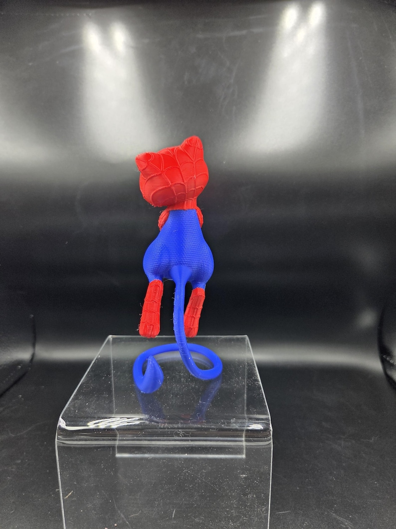 May include: A 3D-printed figurine of a cat in a Spider-Man costume. The cat has a red head and paws, a blue body, and a curled blue tail. The figurine is displayed on a clear, rectangular stand.