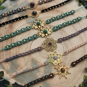 Puede incluir: Una colección de pulseras de macramé hechas a mano con diseños intrincados. Las pulseras presentan varios colores, incluyendo verde azulado, negro y marrón, y están adornadas con detalles metálicos dorados y piedras preciosas. Las pulseras se exhiben sobre una superficie de madera.