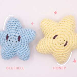May include: Two crocheted star-shaped plushies, one light blue labeled BLUEBELL and one pale yellow labeled HONEY. Each star features dark brown embroidered eyes and a simple stitched mouth, with small pink star icons surrounding the items.