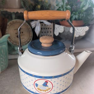 May include: White enamel tea kettle with a blue lid and a wooden handle. The kettle features a blue and white floral pattern with a chicken illustration. The spout is white, and the handle is made of light-colored wood.